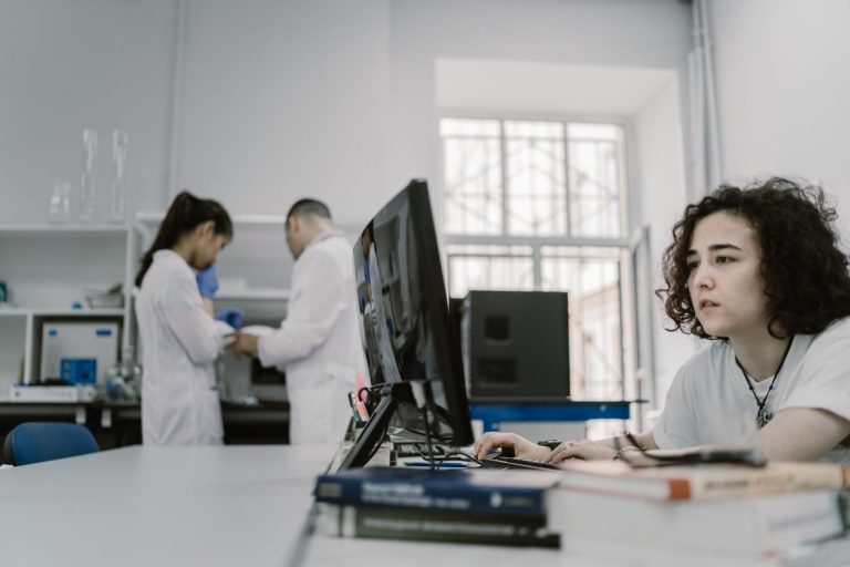 Pesquisadora trabalha em um computador dentro de um laboratório, em meio a equipamentos e atividades de pesquisa.